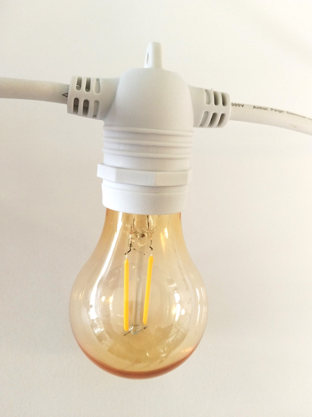 A clear glass LED light bulb with a warm amber tint, suspended in a white socket with a white power cord visible at the top and sides.