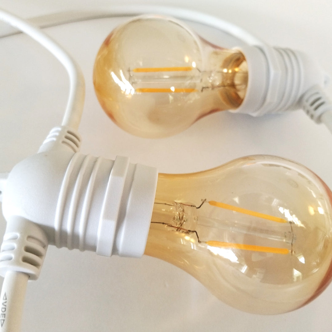 Two clear glass globe string lights with warm amber-tinted bulbs and white screw-in sockets on white cords, photographed close up on a light background.