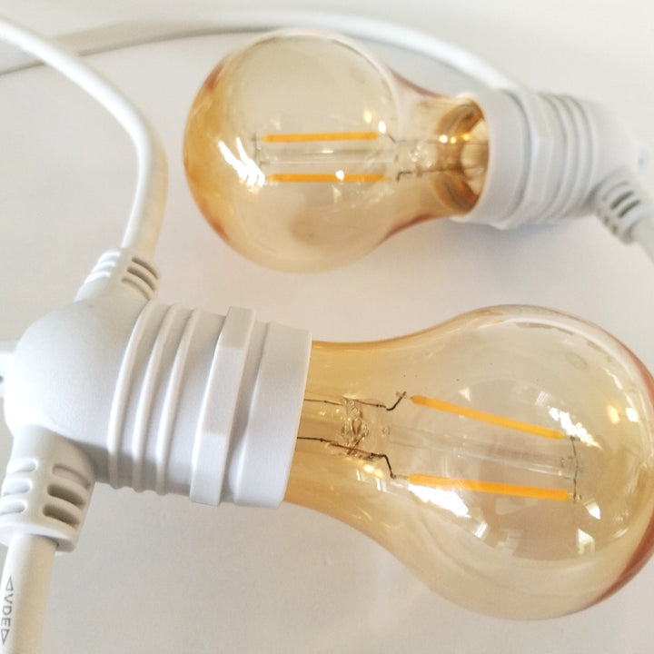 Two clear glass globe string lights with warm amber-tinted bulbs and white screw-in sockets on white cords, photographed close up on a light background.