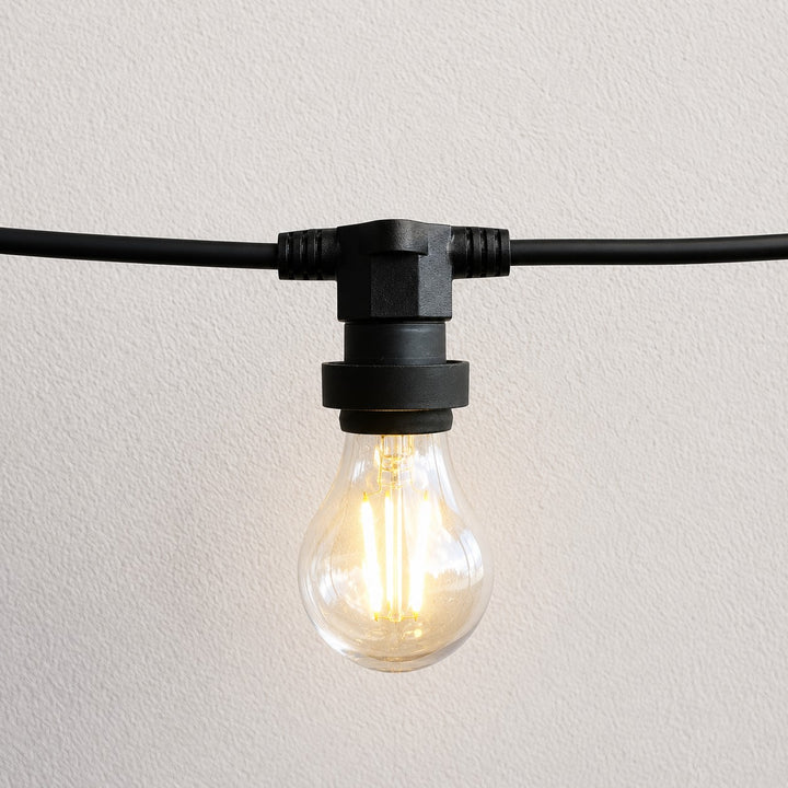 A single black outdoor-style light fixture with a clear glass globe bulb glowing warm yellow, mounted on a black cable against a light textured wall.
