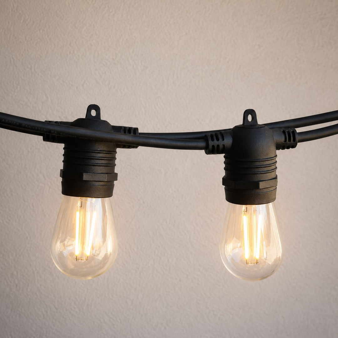 Black string light bulbs with clear glass and visible warm glowing filaments, mounted on a black cable against a light beige textured wall.